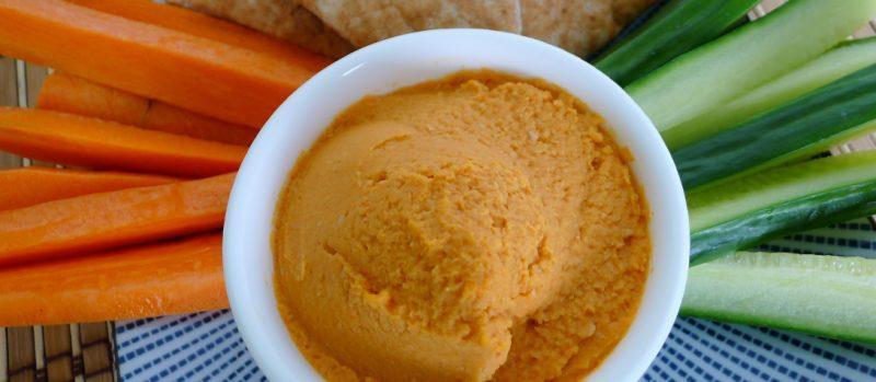 white bowl of red pepper hummus and carrot + cucumber sticks
