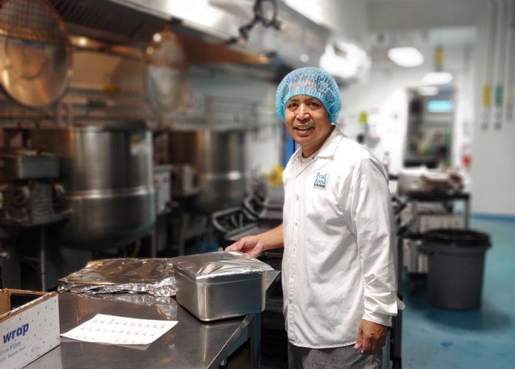 man in kitchen facility wearing white coat, blue hairnet, smiling