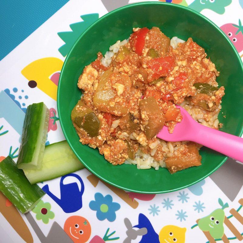 green bowl with ratatouille and rice with pink spoon, kids placemat and cucumber sticks in background
