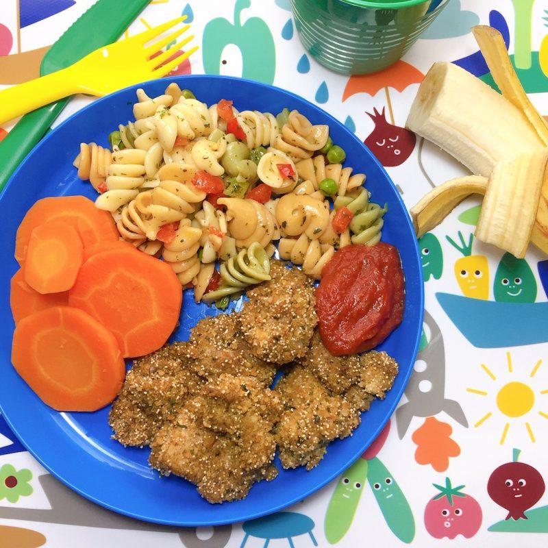 blue plate with carrots, chicken meteorites, ketchup, pasta salad, background of kids placemat and peeled banana