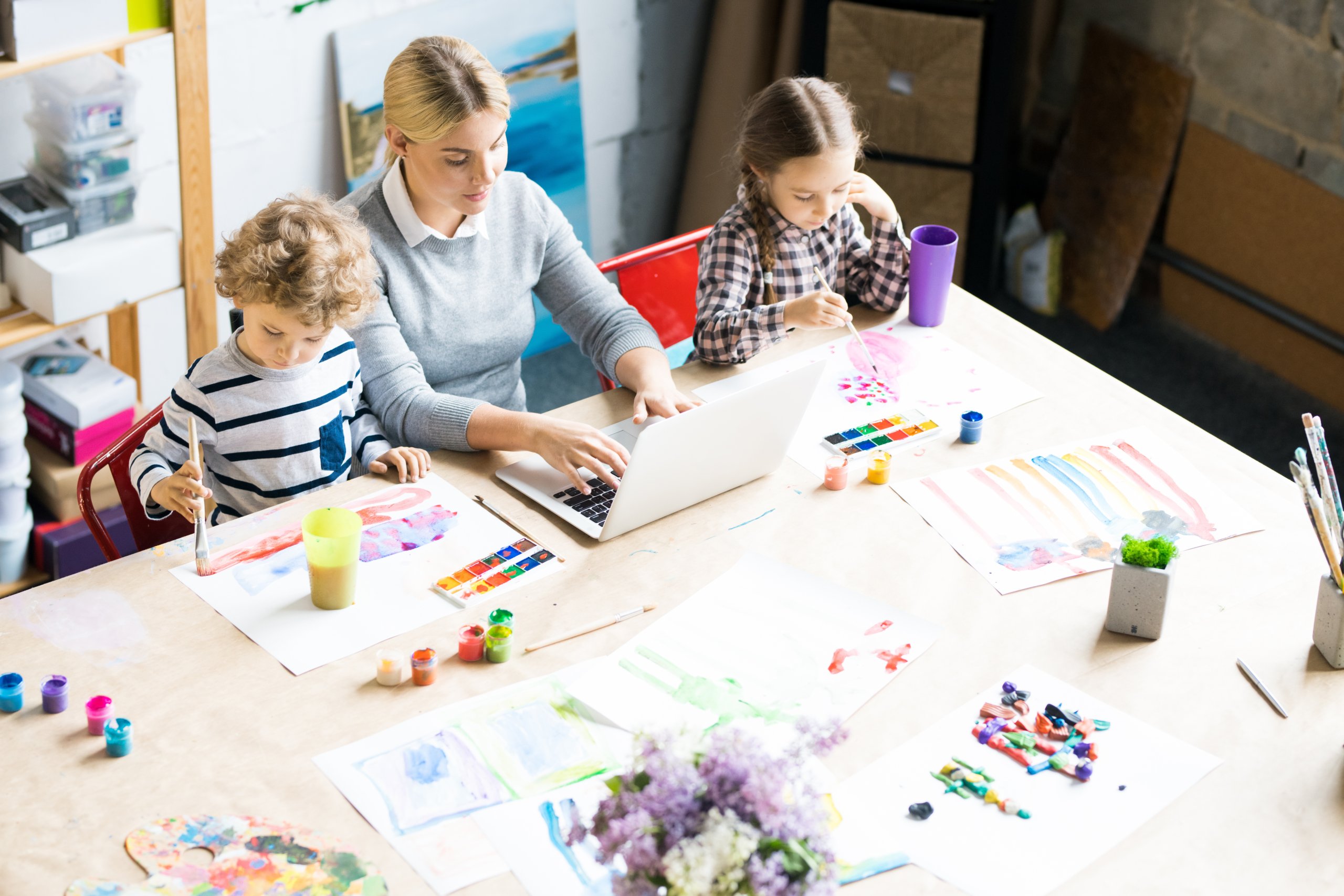 High angle portrait of young woman working at laptop with two kids painting beside her at table in modern apartment