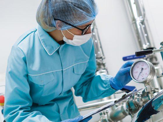 lab tech woman wearing glasses, mask, hairnet, blue jacket with clipboard