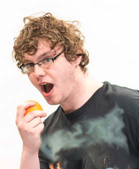 curly haired man in black t shirt about to bite into an orange