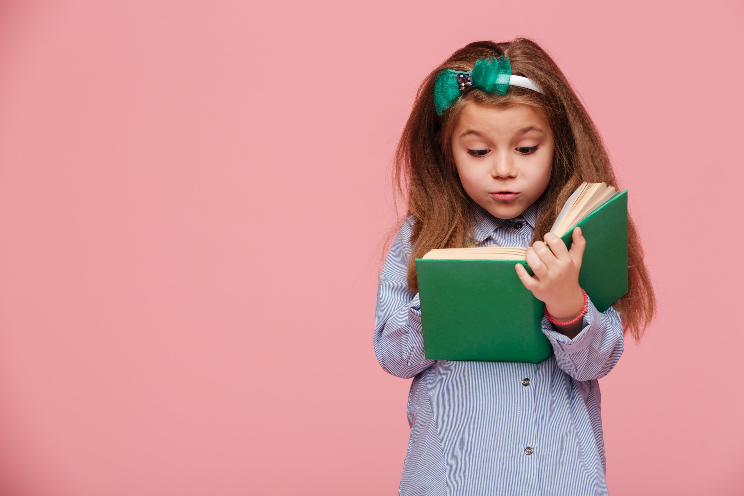Image of cute girl 5-6 years with long auburn hair reading interesting book, being involved in education isolated over pink background