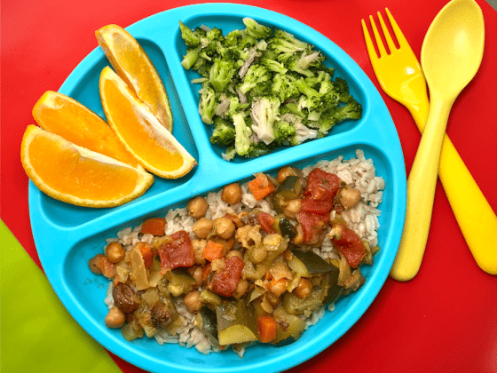a child's blue divided plate with garbanzo bean tajine, brown rice, steamed broccoli and orange slices next to yellow spoon and fork
