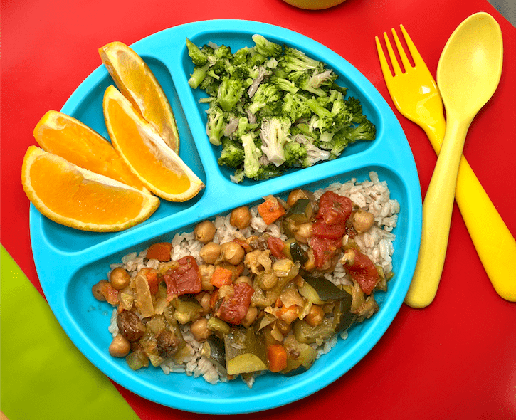 a child's blue divided plate with garbanzo bean tajine, brown rice, steamed broccoli and orange slices next to yellow spoon and fork