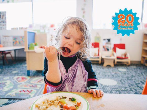 little girl with blonde hair happily eating lunch at child care
