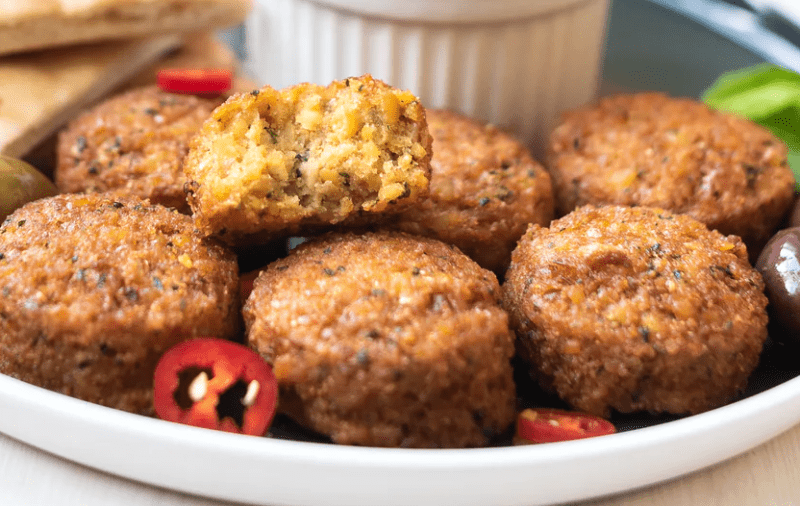 Mini falafels arranged on a dish in front of a ramekin with scattered sliced peppers