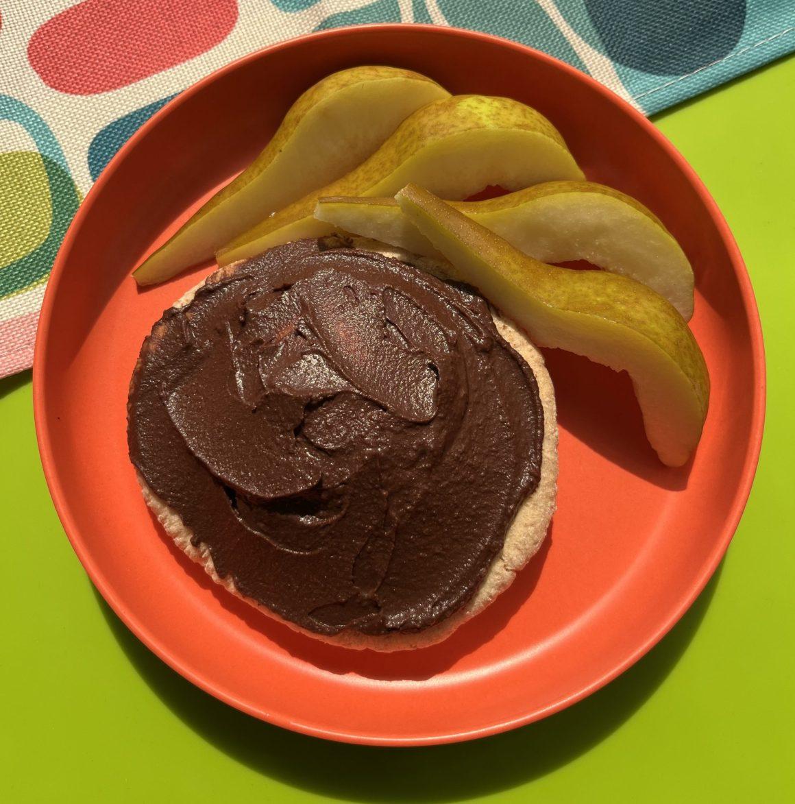 RFRK's cocoa chic'pea spread on top of whole white pita next to slices of fresh pear on a green table and red plate