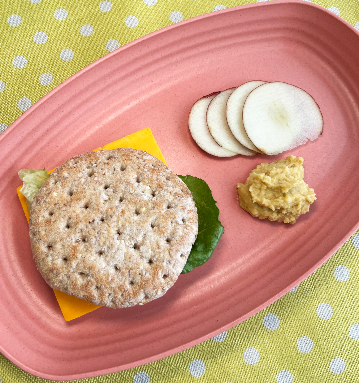 whole white pita sandwich with hummus and lettuce with sliced apples and hummus on the side on a pink plate and yellowish green dotted placement