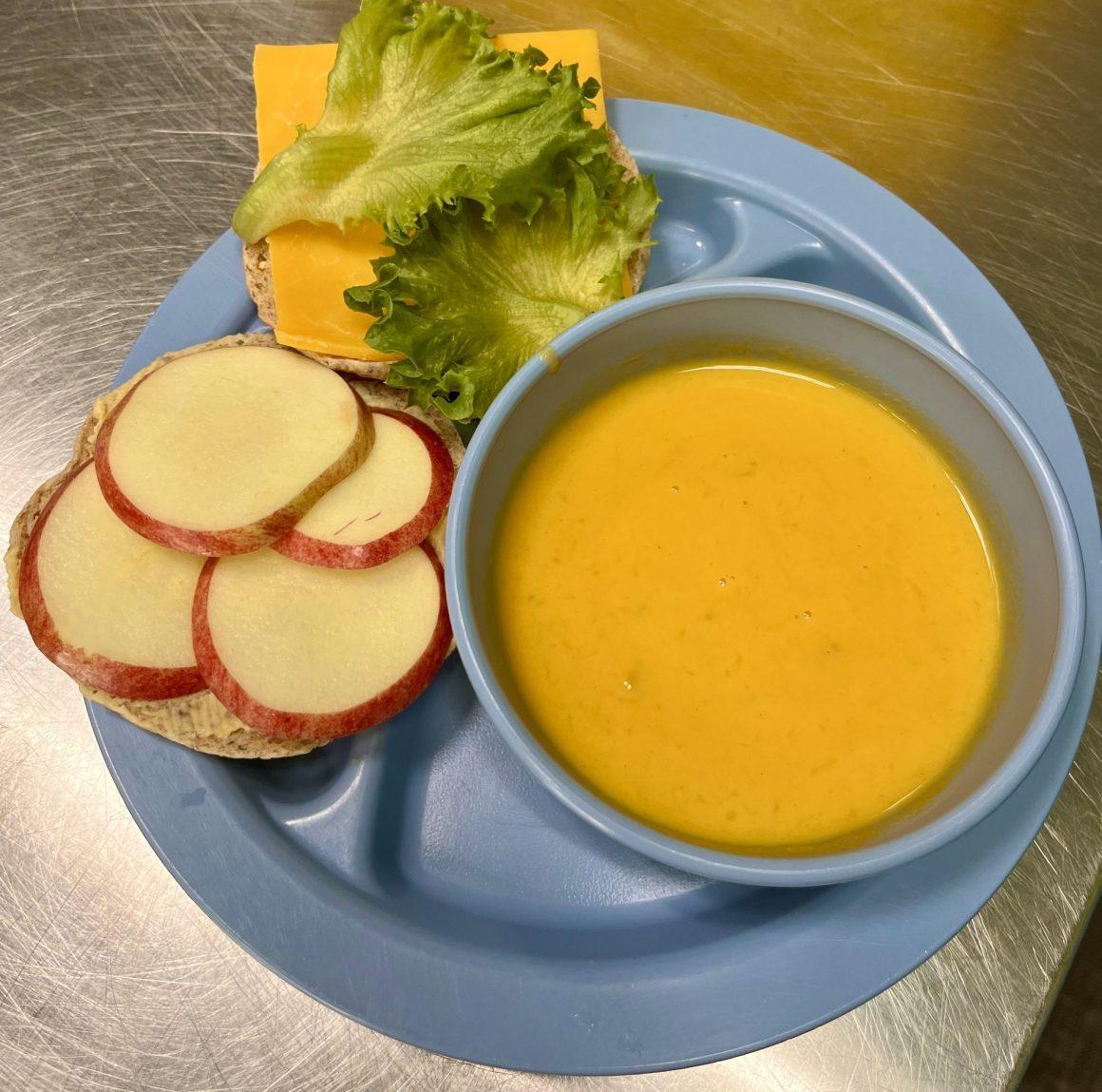 hummus cheddar apple sandwich on open whole wheat pita next to bowl of coconut squash soup on a blue plate