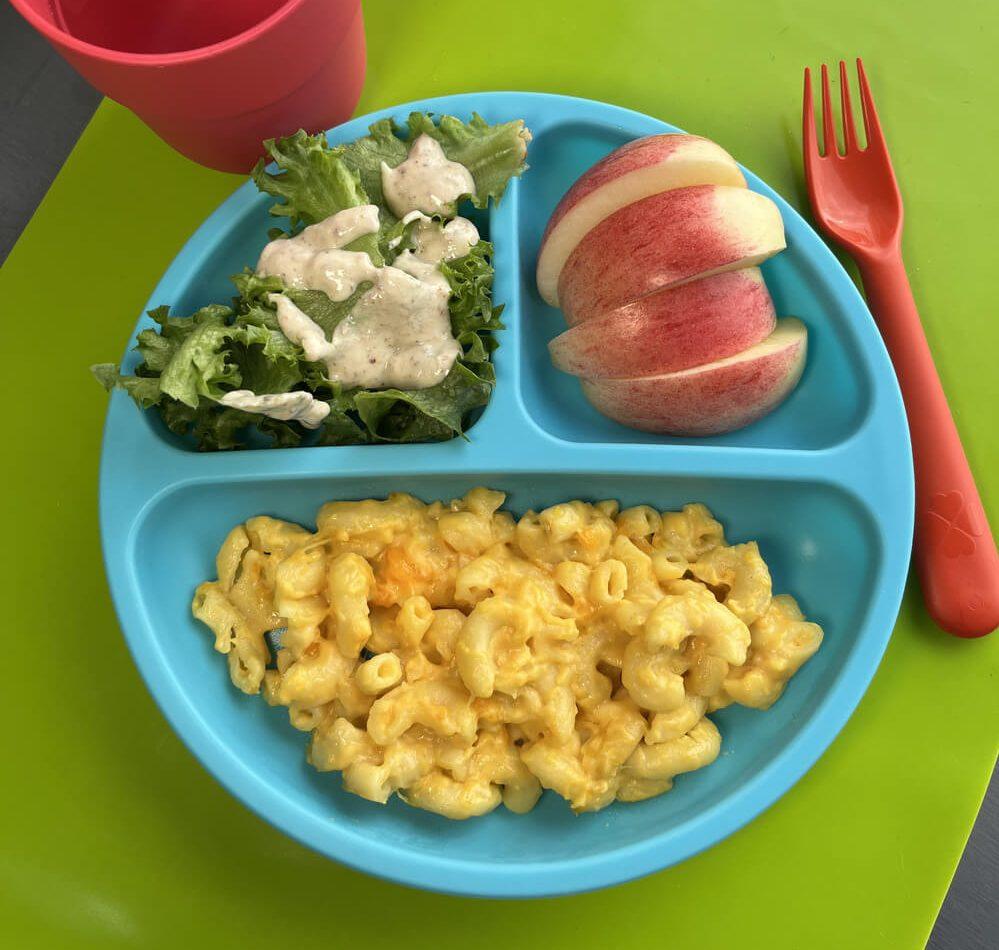 blue kids plate of RFRK mac 'n cheese with side caesar salad with homemade RFRK caesar dressing and sliced apple. Red fork, green placement and red cup