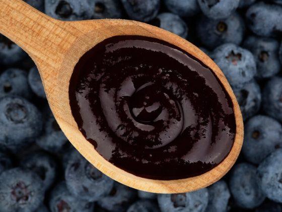 close up of blueberry puree in a wooden spoon in front of a background of whole fresh blueberries