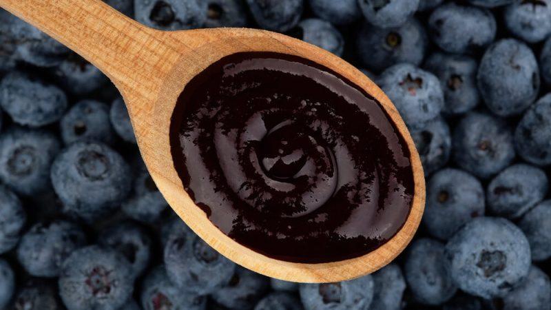 close up of blueberry puree in a wooden spoon in front of a background of whole fresh blueberries