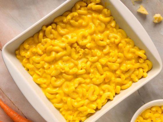 a white baking and serving dish filled with mac n cheese surrounded by cheese and carrots on a table