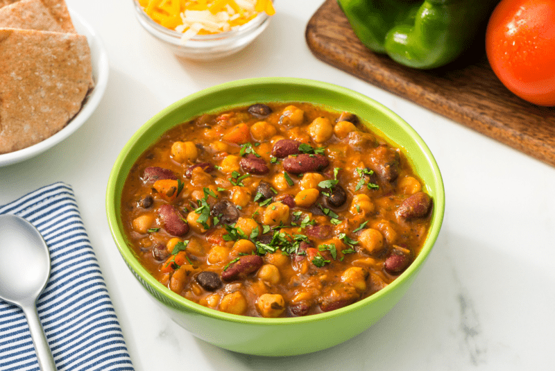 RFRK's chili chili bang bang in a green bowl served on a table next to a spoon and corner view of a tomato and green pepper