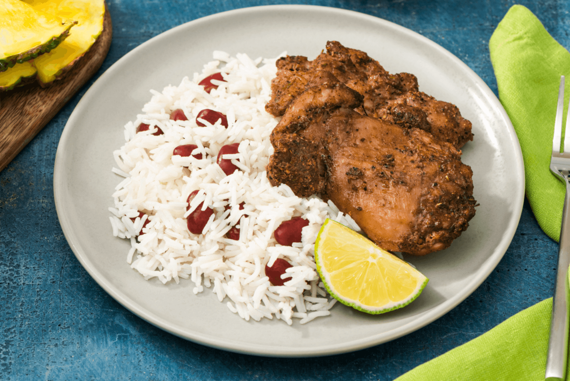 A plate of RFRK's jerk chicken served with beans and rice and a lime wedge on a white plate and blue table