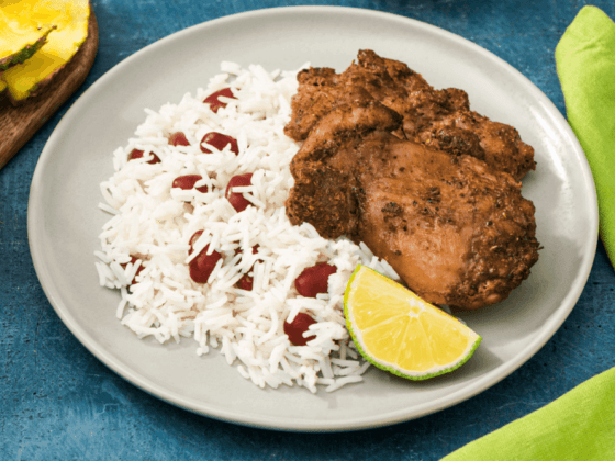 a plate of jerk chicken next to rice and beans and slice of lime on white plate on blue table