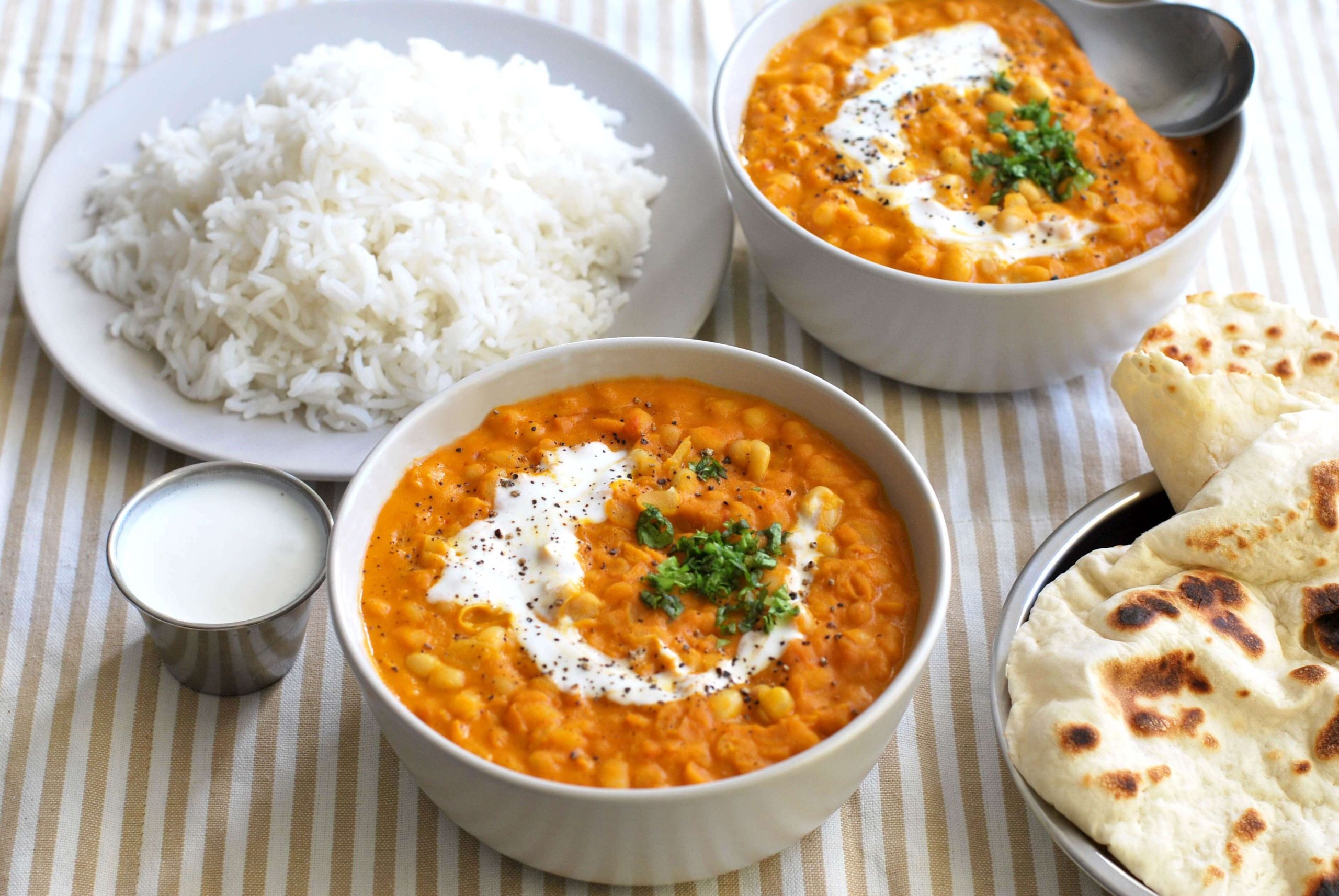 Two white bowls of white bean curry with one plate of rice next to it and a portion of a photo of naan in the lower right corner
