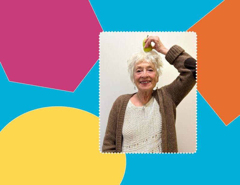 lady with white shirt and brown sweater smiling holding pear on top of head in front of multi colour background of purple, blue, orange and yellow