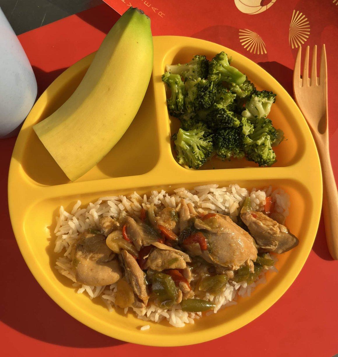 yellow kids plate on red placement with yellow fork with half banana, broccoli, and gong bao chicken served over basmati rice