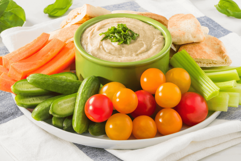 green ramekin filled with rfrk's beany basil dip surrounded by veggies including carrots, baby tomatoes, celery and pita