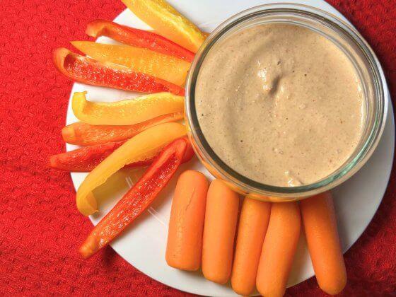 small bowl of red pepper saucy dipz with carrots and red peppers on white plate with red background