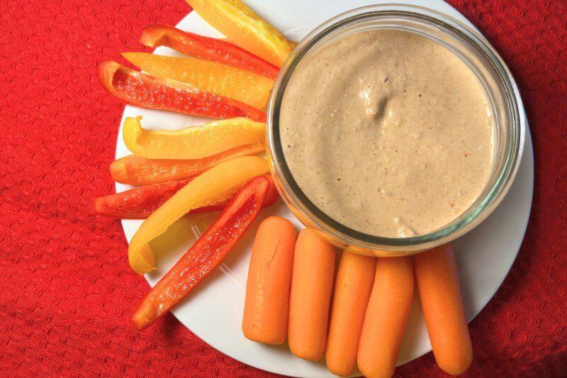 small bowl of red pepper saucy dipz with carrots and red peppers on white plate with red background