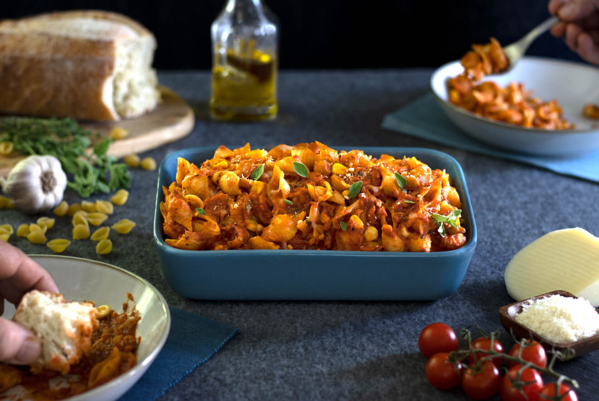 RFRK's vegetarian pasta bake in a large blue casserole style dish with bread, tomatoes, olive oil, cheese scattered around main casserole dish