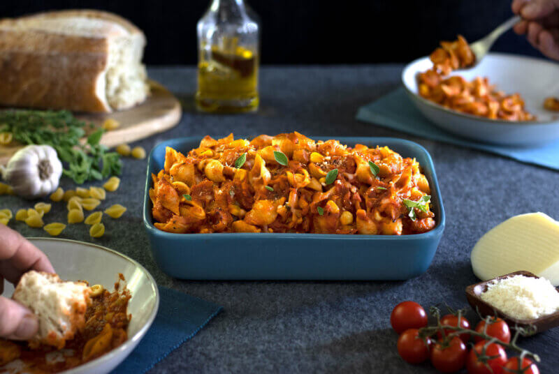 RFRK's vegetarian pasta bake in a large blue casserole style dish with bread, tomatoes, olive oil, cheese scattered around main casserole dish