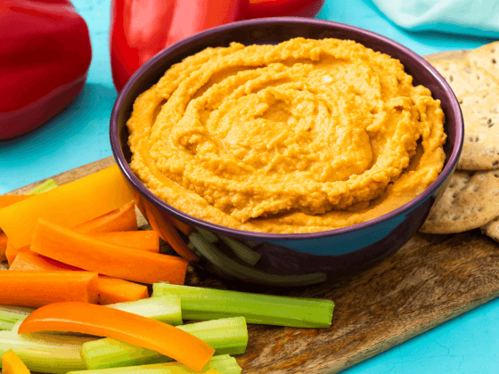 rfrk's red pepper hummus in small black bowl next to veggie sticks, carrot sticks and crackers