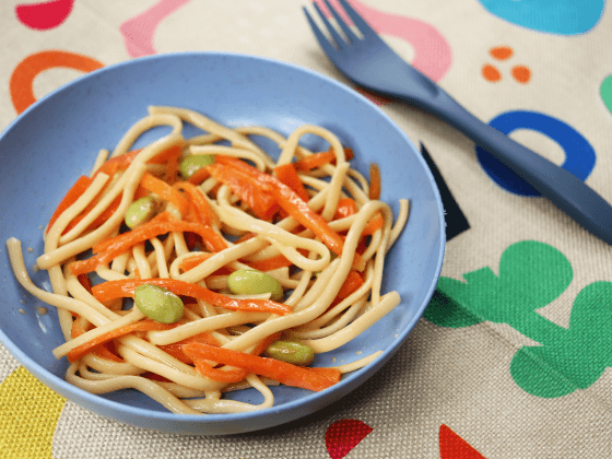 close up of ginger-soy noodles w/ edamame snack in light blue bowl next to dark blue fork on top of colourful shapes place mat