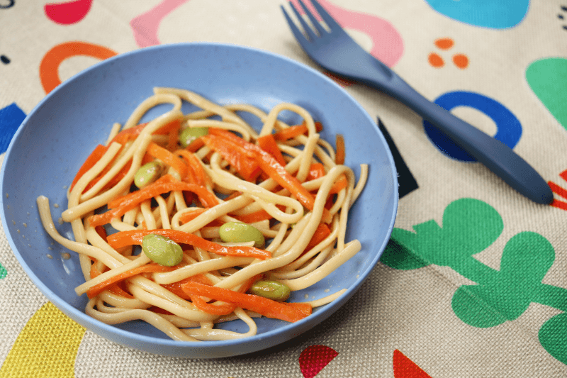 close up of ginger-soy noodles w/ edamame snack in light blue bowl next to dark blue fork on top of colourful shapes place mat