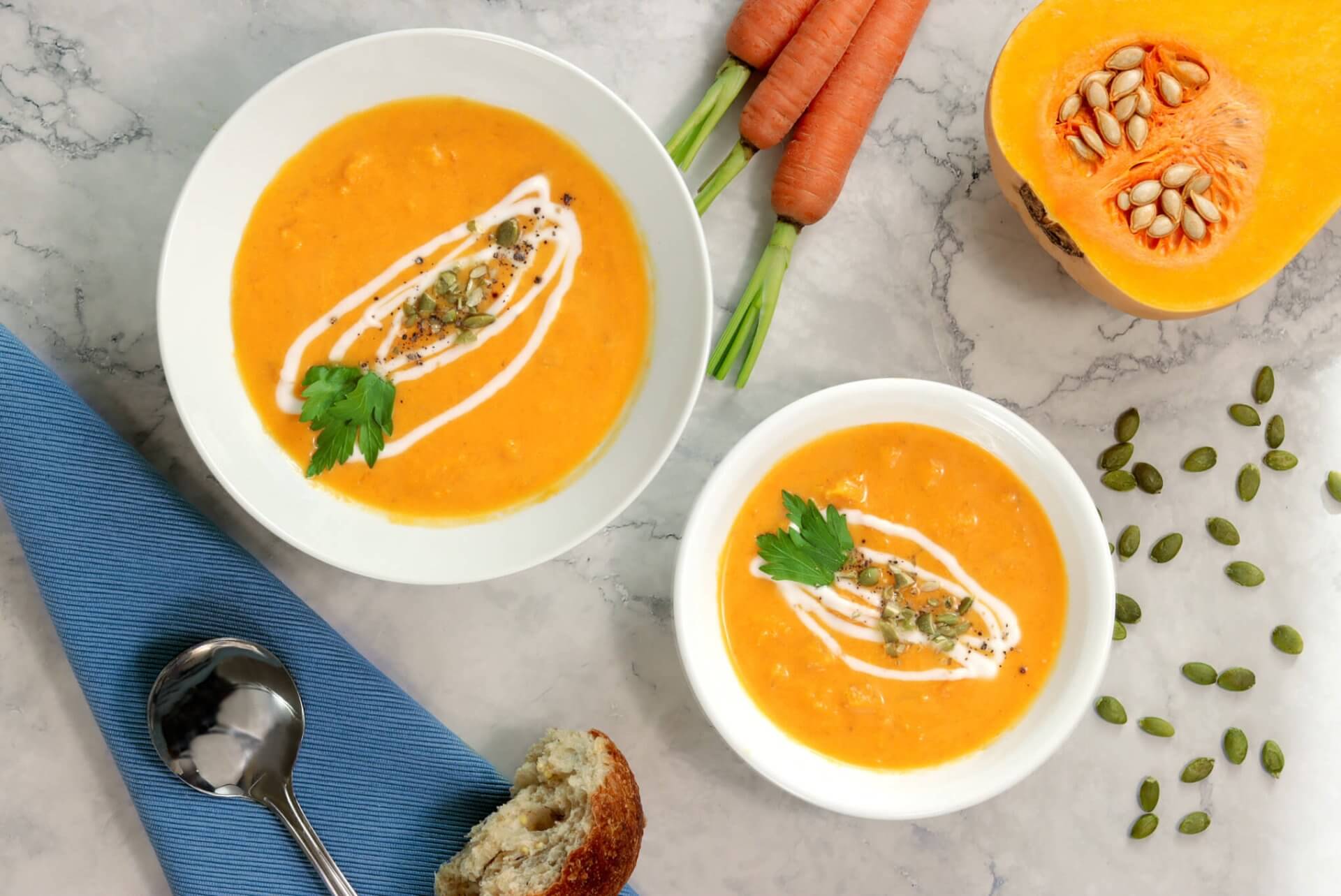 two bowls of rfrk's squash and coconut soup with garnish next to sliced open butternut squash and carrot