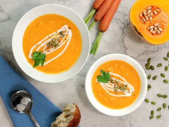 two bowls of rfrk's squash and coconut soup with garnish next to sliced open butternut squash and carrot