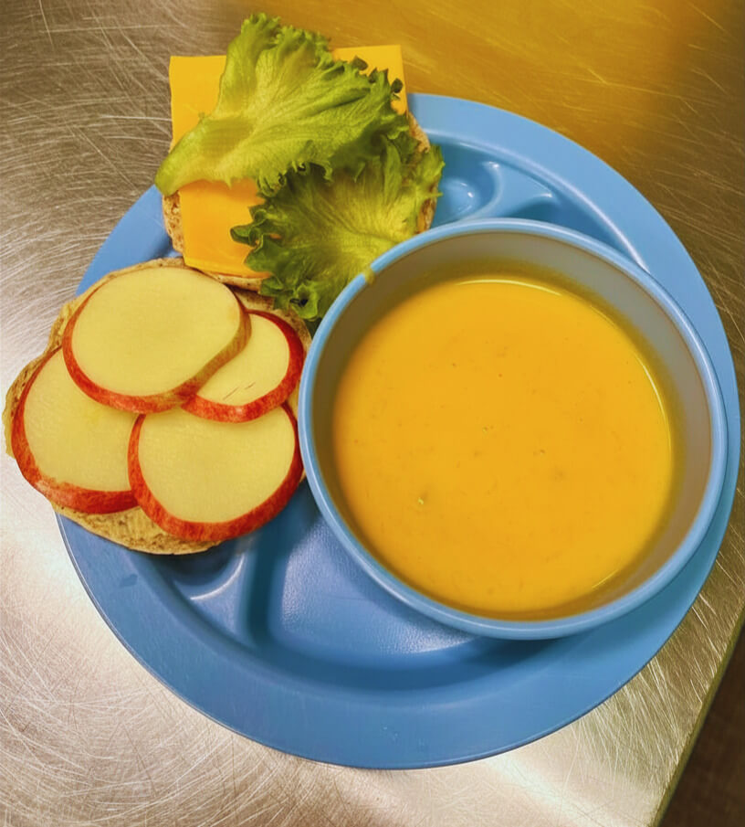 blue plate with blue bowl of rfrk's squash and coconut soup with sandwich of sliced apple, hummus and cheddar cheese