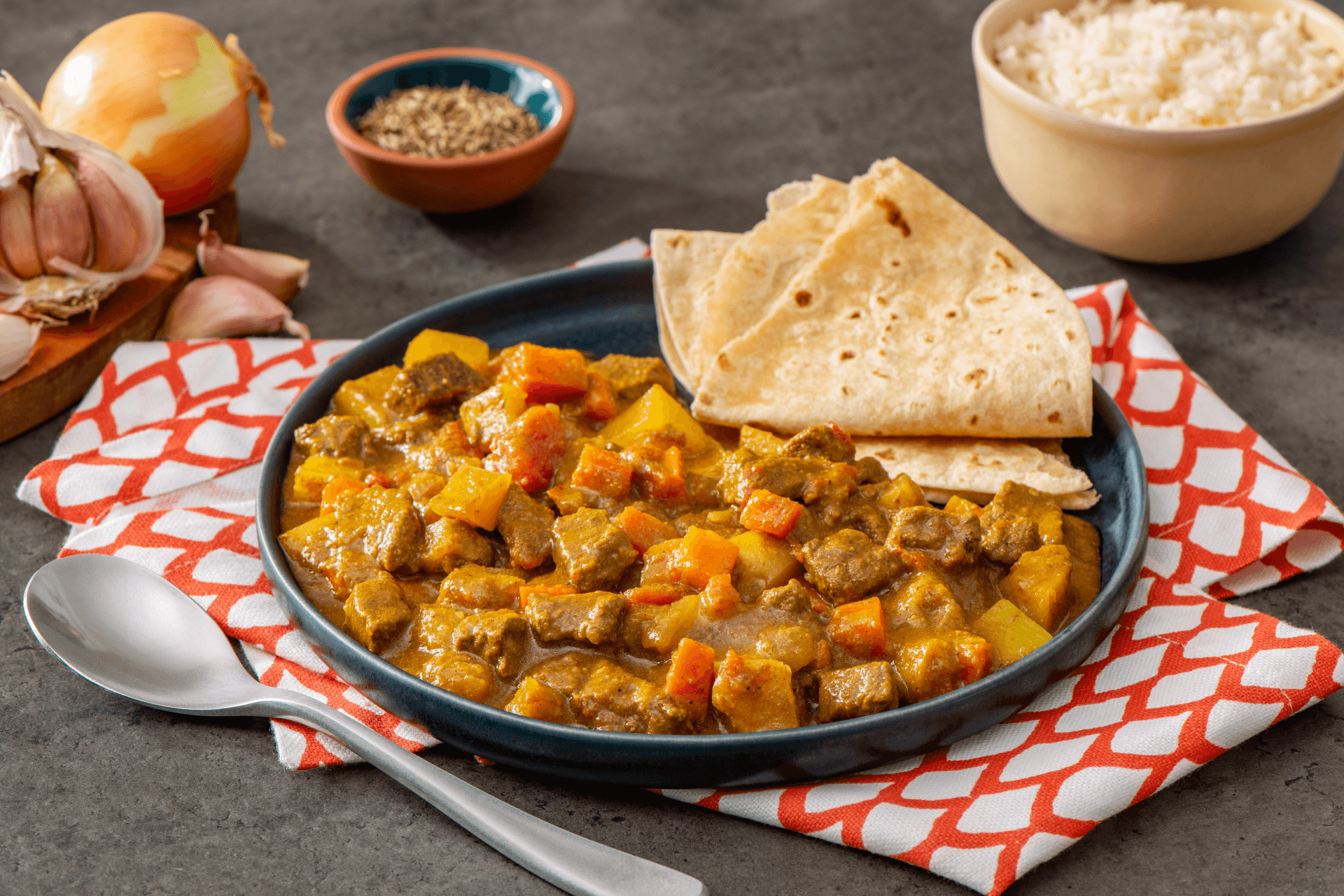 large black plate with trinidadian curry beef served next to folded naan surrounded by accompaniments and red and white napkins