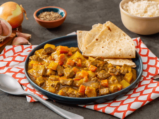 large black plate with trinidadian curry beef served next to folded naan surrounded by accompaniments and red and white napkins