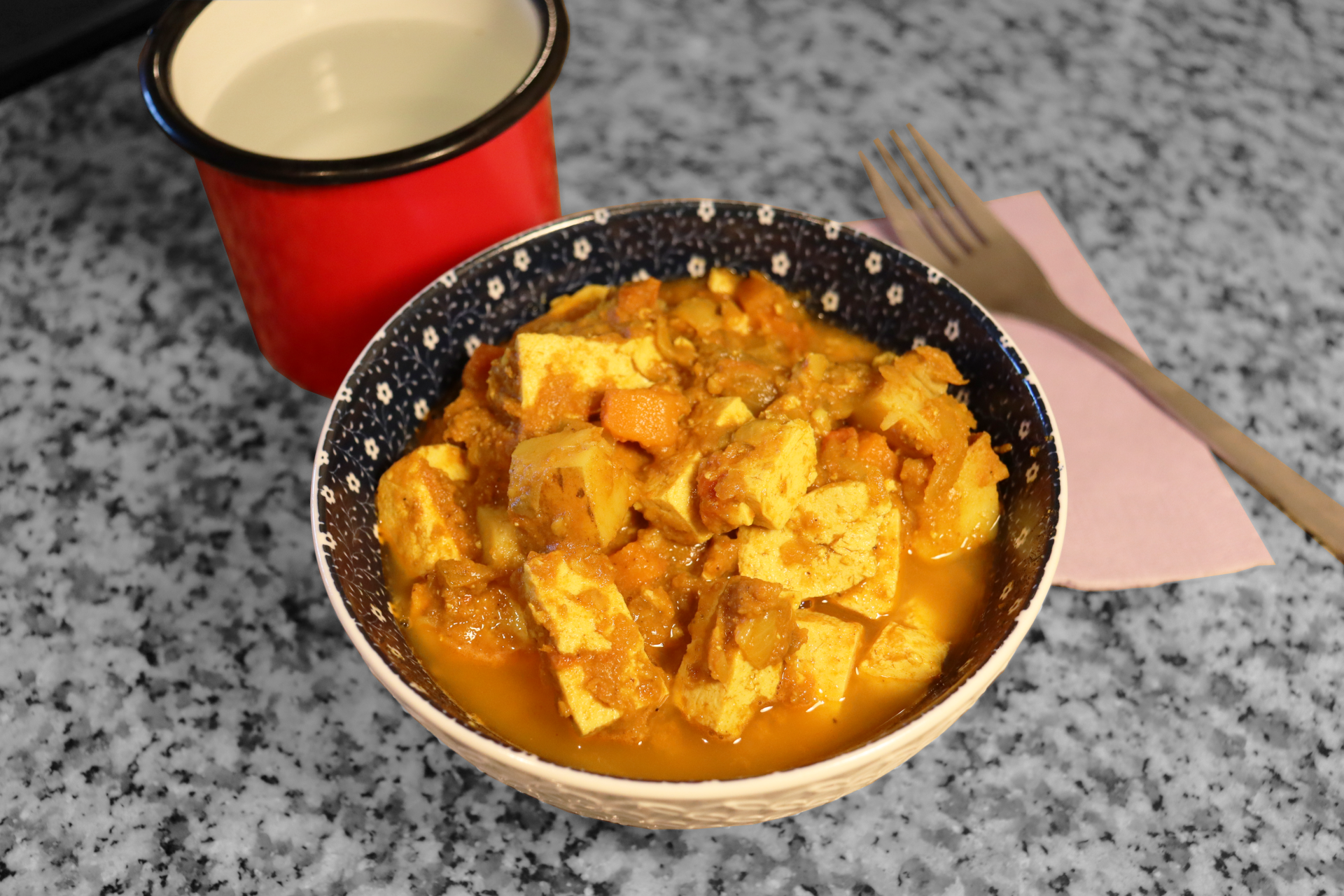 bowl of trinidadian curry tofu next to fork on pink napkin on top of countertop next to red cup