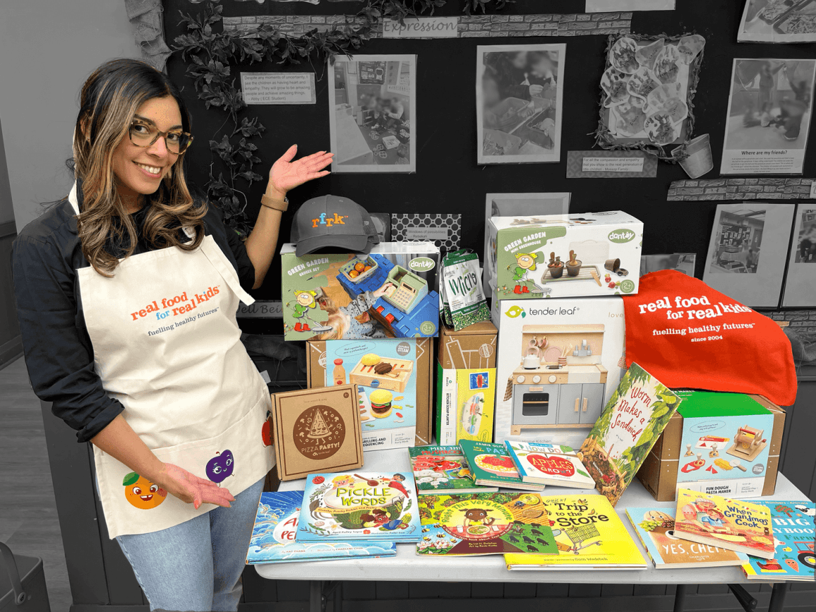 grand prize drop off! 1 rfrk client experience coordinator leticia pictured next to large grand prize package filled with kids books, kids toys, rfrk swag and other gifts