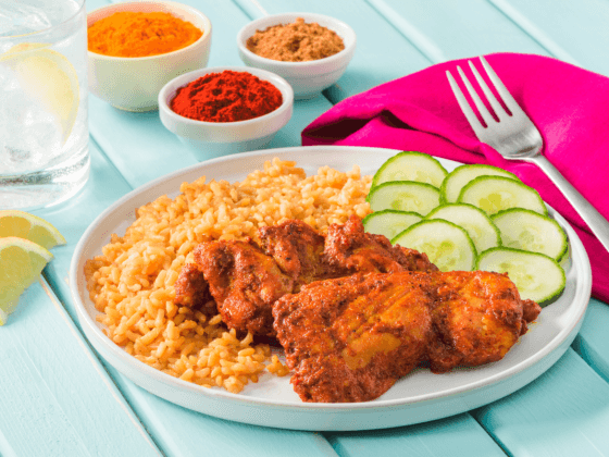 mumbai chicken on plate served with sliced cucumber next to fork on bright reddish pink napkin and small dishes with spices