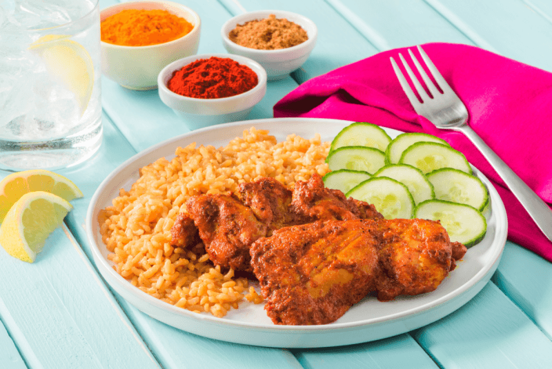mumbai chicken on plate served with sliced cucumber next to fork on bright reddish pink napkin and small dishes with spices
