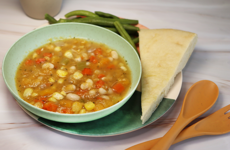 bowl of three sisters soup with beans, squash and corn served in light green bowl next to bread and green beans